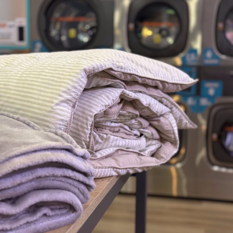 Lavandería de Autoservicio en Fuengirola Mantas enrolladas sobre una mesa, con lavadoras de autoservicio de fondo.